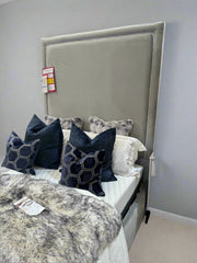 A double bed base and mattress with a 74-inch tall headboard upholstered in grey velvet with white piping, accompanied by decorative pillows, in a furniture showroom setting.