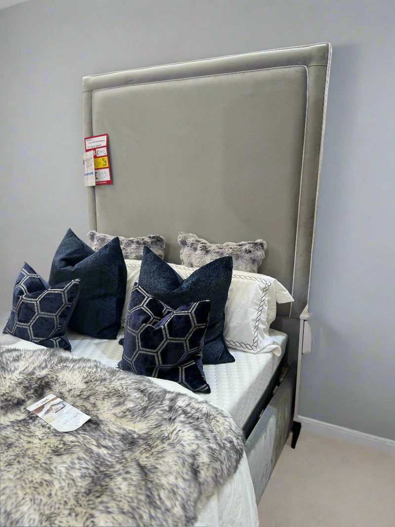 A double bed base and mattress with a 74-inch tall headboard upholstered in grey velvet with white piping, accompanied by decorative pillows, in a furniture showroom setting.